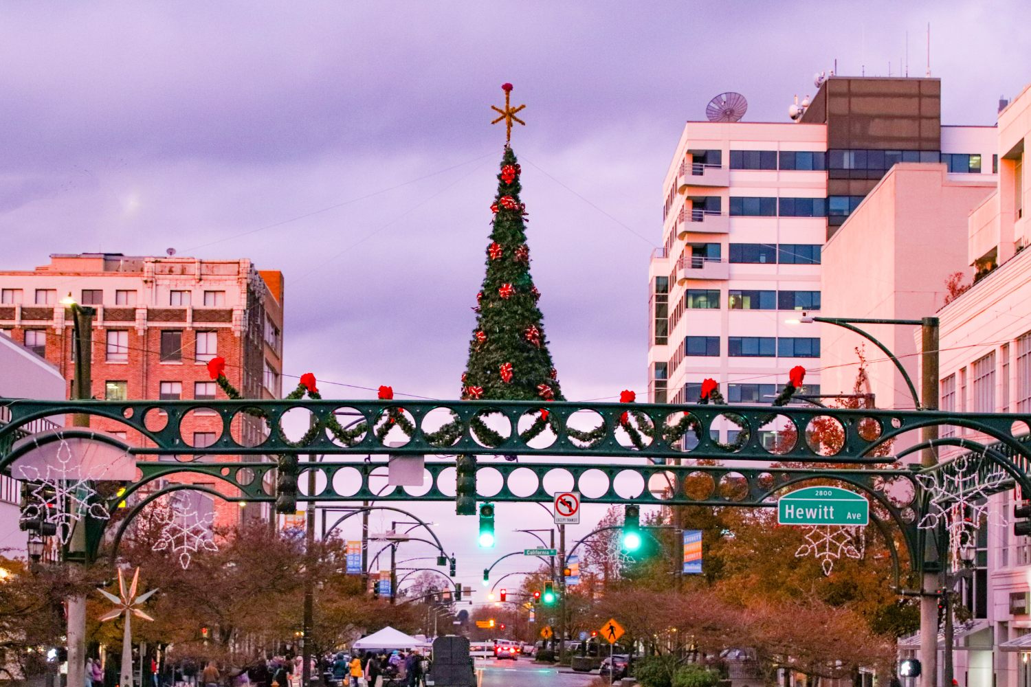 A festive holiday scene in downtown Everett features a large decorated Christmas tree, colorful lights, and seasonal decorations along Hewitt Avenue under a cloudy evening sky.