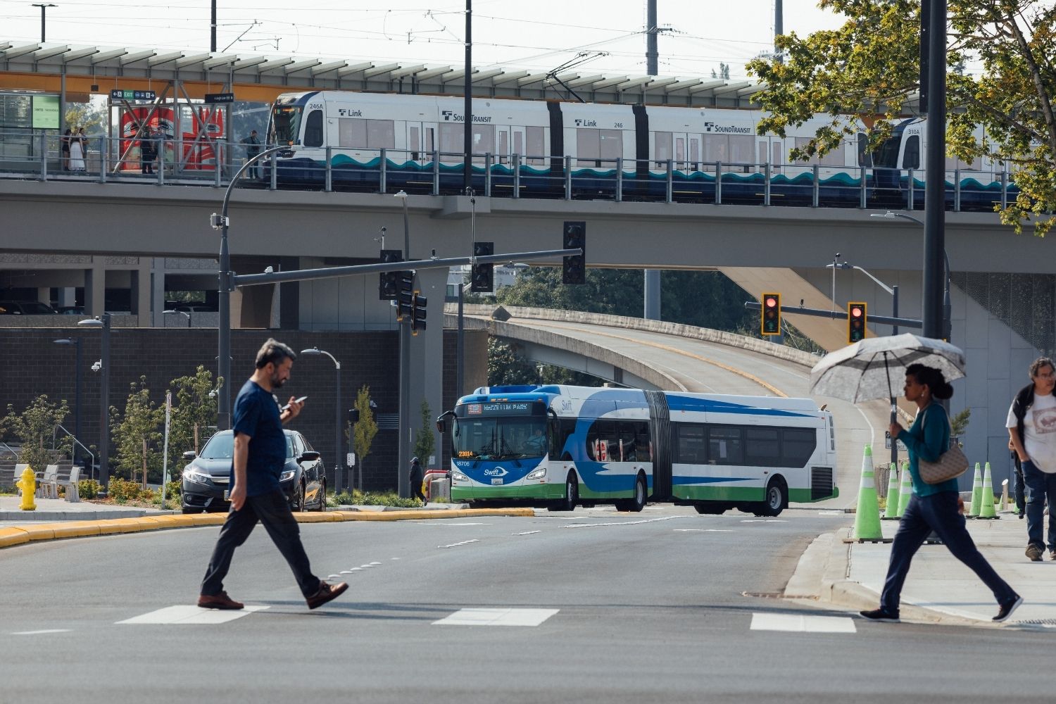 People crossing the street at Lynnwood City Center Station. In the background is a Swift Orange Line bus and the Link light rail.