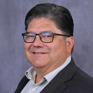 A professional headshot of a man wearing glasses and a dark blazer over a light-colored checkered shirt. He has short, dark hair and is smiling, posed in front of a neutral gray background.