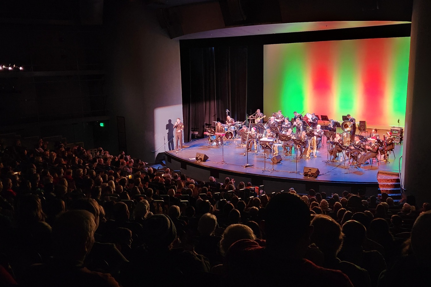 A full audience watches a live performance by a large band on a brightly lit stage with a red and green backdrop.