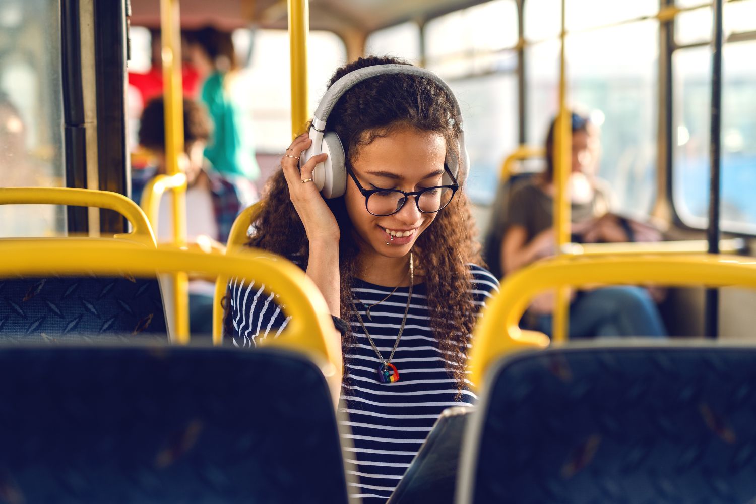 A young woman with curly hair and a lip piercing sits on the bus. She is wearing a striped shirt and has headphones on. She is looking down at her phone and smiling.