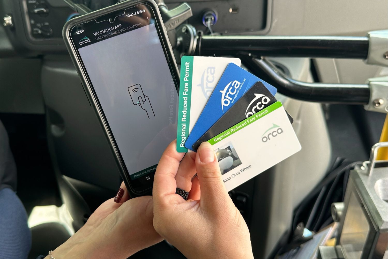 A hand holding four different types of ORCA cards in front of a portable ORCA card scanner. The background is the inside of a DART paratransit bus.