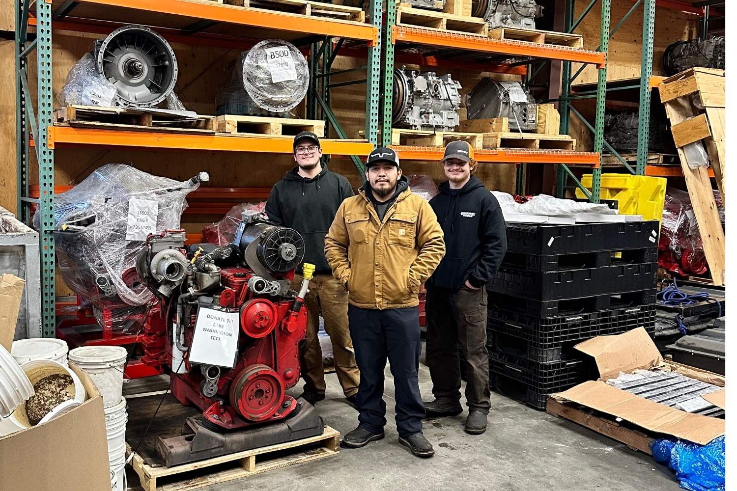 Three people stand in an auto shop in front of an engine and a transmission. 