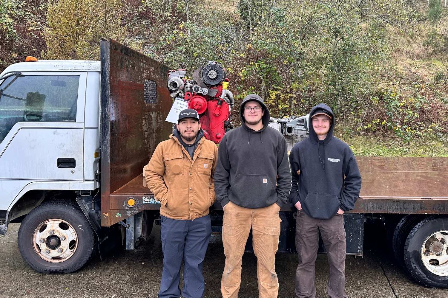 Three people stand in front of a flat bed truck. On the back of the truck is a bus engine and transmission.