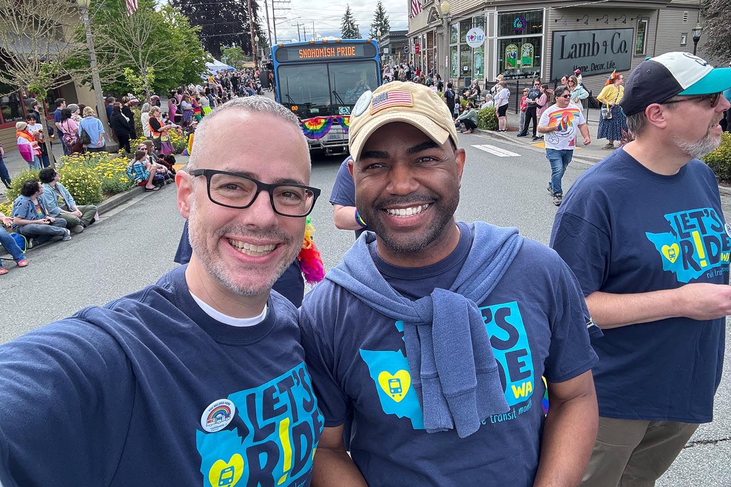 Phillip & Lance smile at the Snohomish Pride celebration. A Community Transit bus decked out with rainbow bunting is shown in the background.
