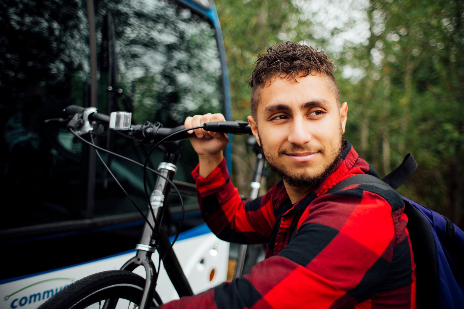 A dark haired man in a red and black plaid shirt looks over his shoulder as he gets his bike off of a bike rack. 