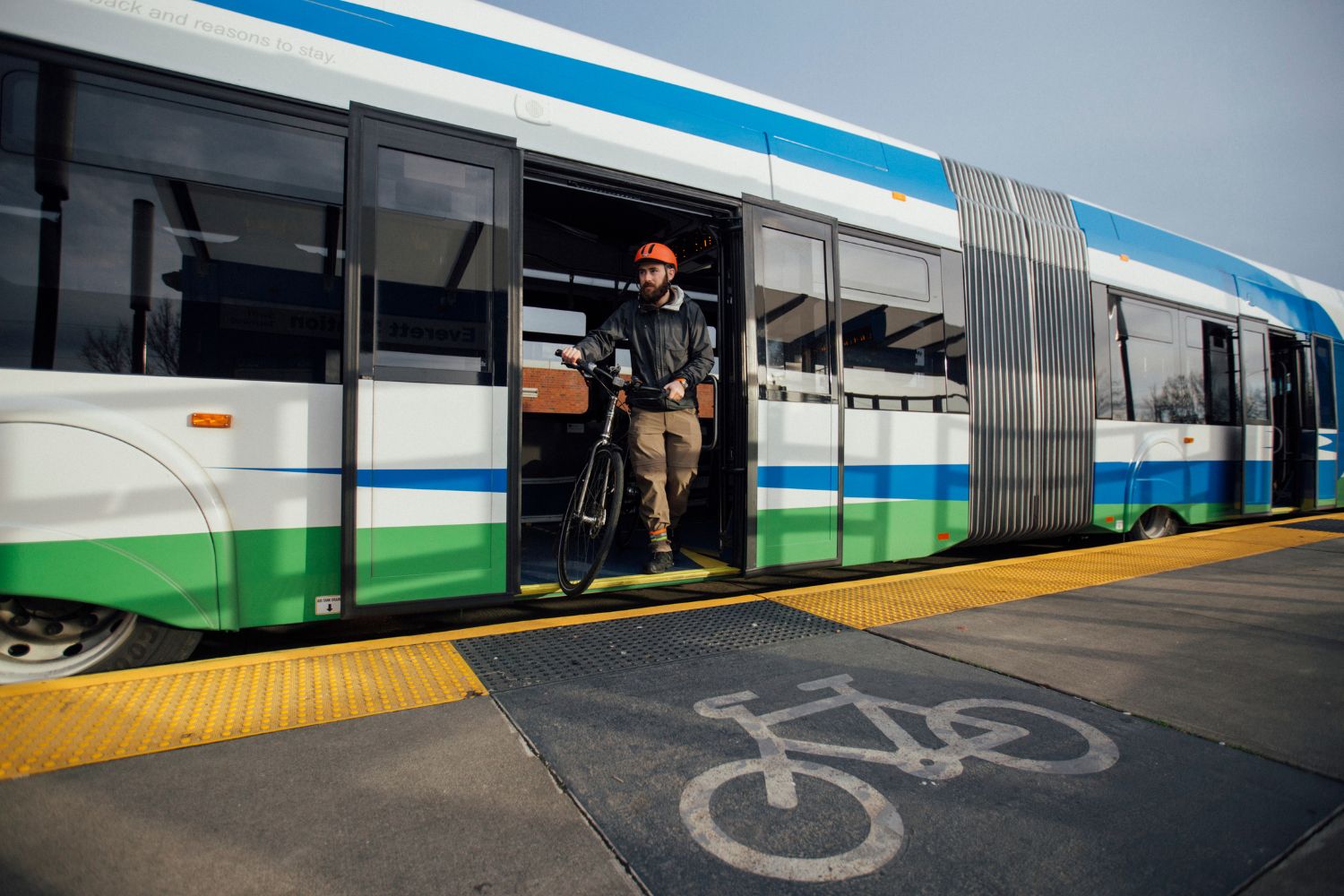 A man with a beard exits a Swift bus. He is pushing his bike off of the bus and looking into the distance. 