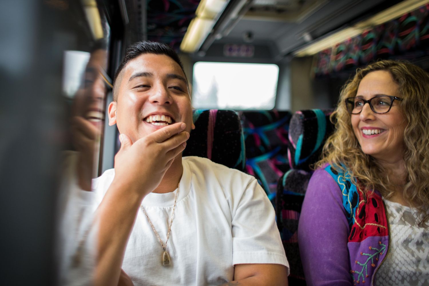 A man in a white shirt and a woman in a purple shirt sit next to each other on the bus. They are both smiling. 