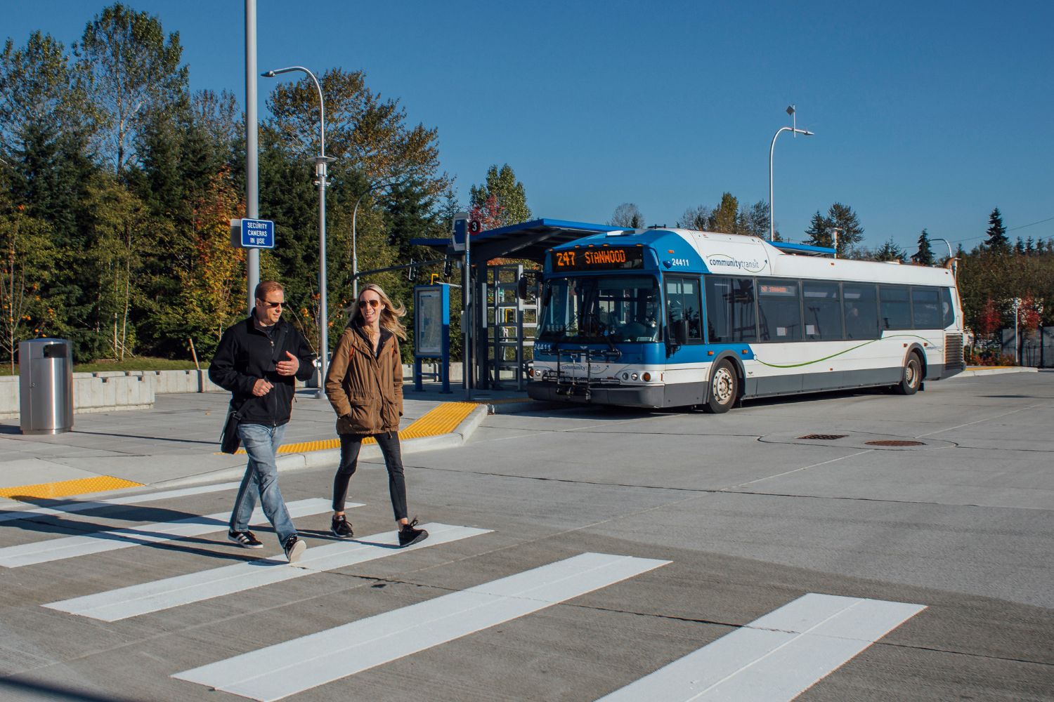 Two people walking together at a transit center on a sunny day. A Route 247 bus is at a bus bay behind them.