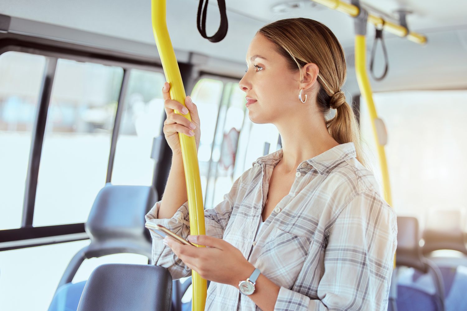 A woman in a plaid shirt is standing on a bus holding on to a yellow pole. She is looking at the window and holding her cell phone in one hand.
