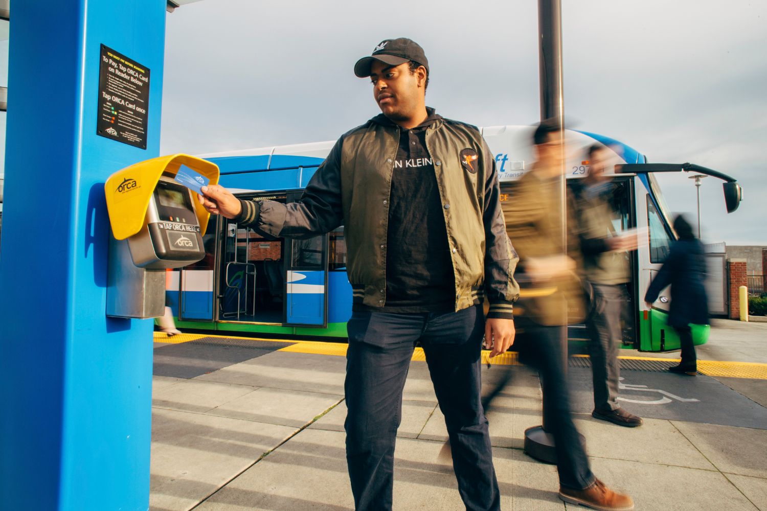 A man standing in front of a Swift bus, scanning his ORCA card on an ORCA card reader.