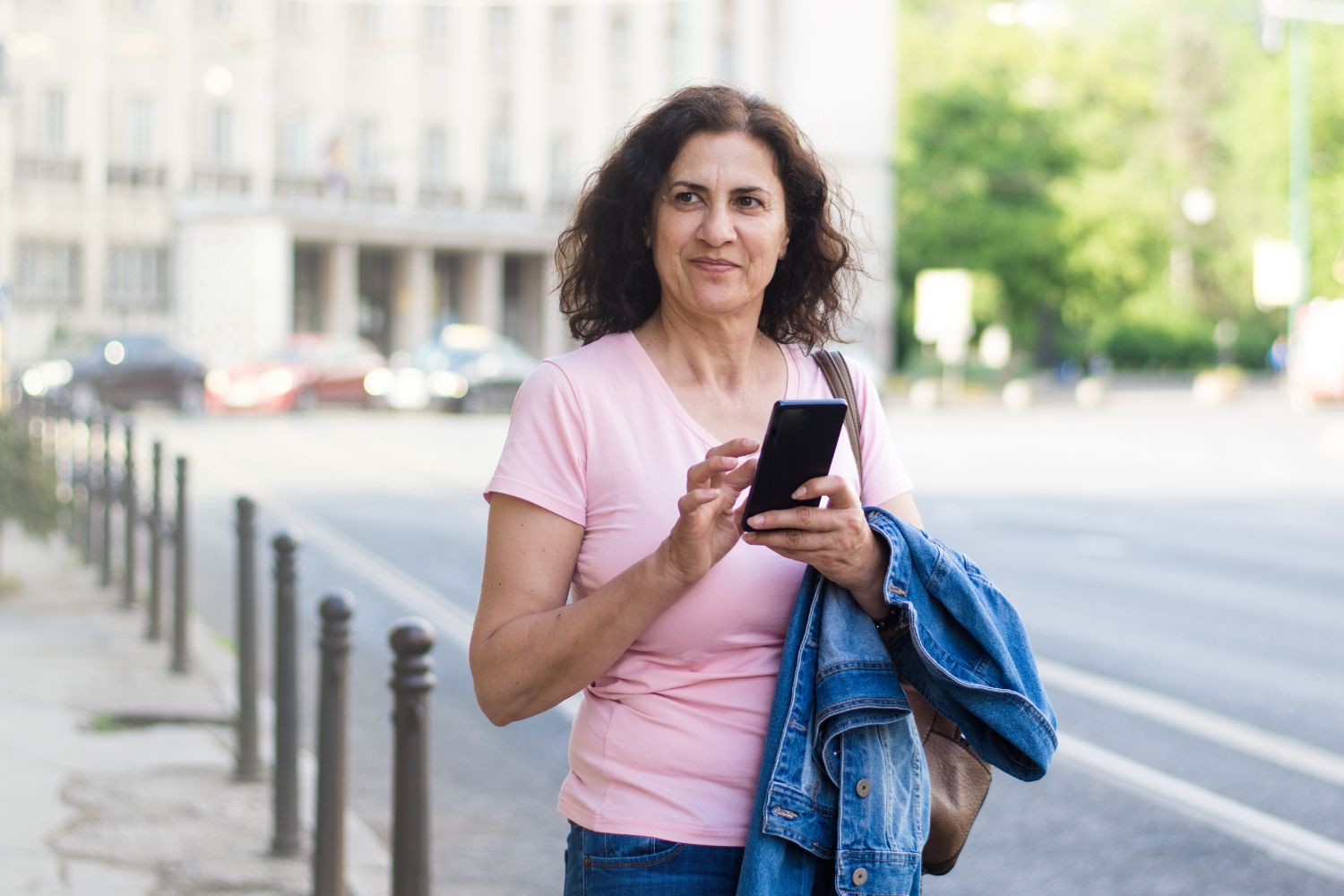 A woman with brown hair and a pink shirt stands in front of a building holding her cell phone. 