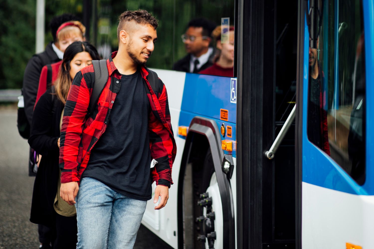 A man in a red flannel walks next to a bus. Behind him are three people waiting to get on the bus. 