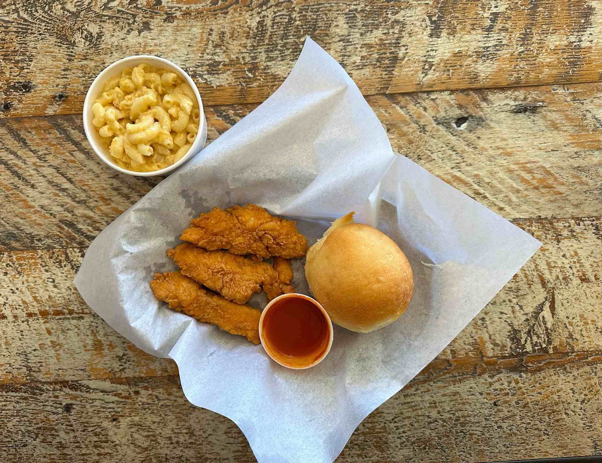 Three chicken tenders with a soft roll on a paper-lined tray. A cup of macaroni and cheese nearby