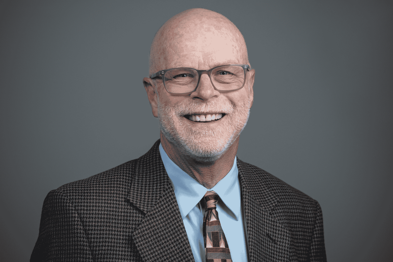 Stanwood Mayor Sid Roberts, wearing glasses, a brown blazer, and a patterned tie, smiles warmly against a neutral gray background.