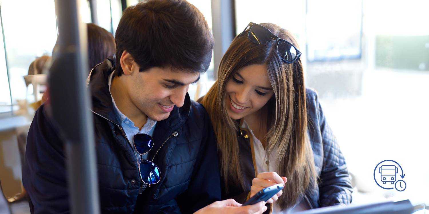 A man and woman sit next to each other on a bus and look at the man's mobile phone