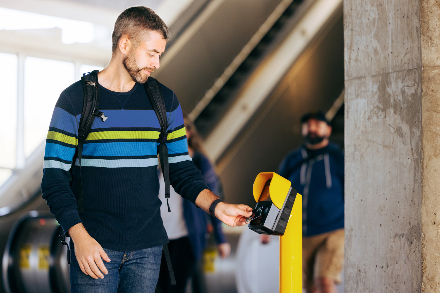 Person tapping an ORCA card at a yellow fare reader in a transit station, with escalators and other commuters visible in the background.