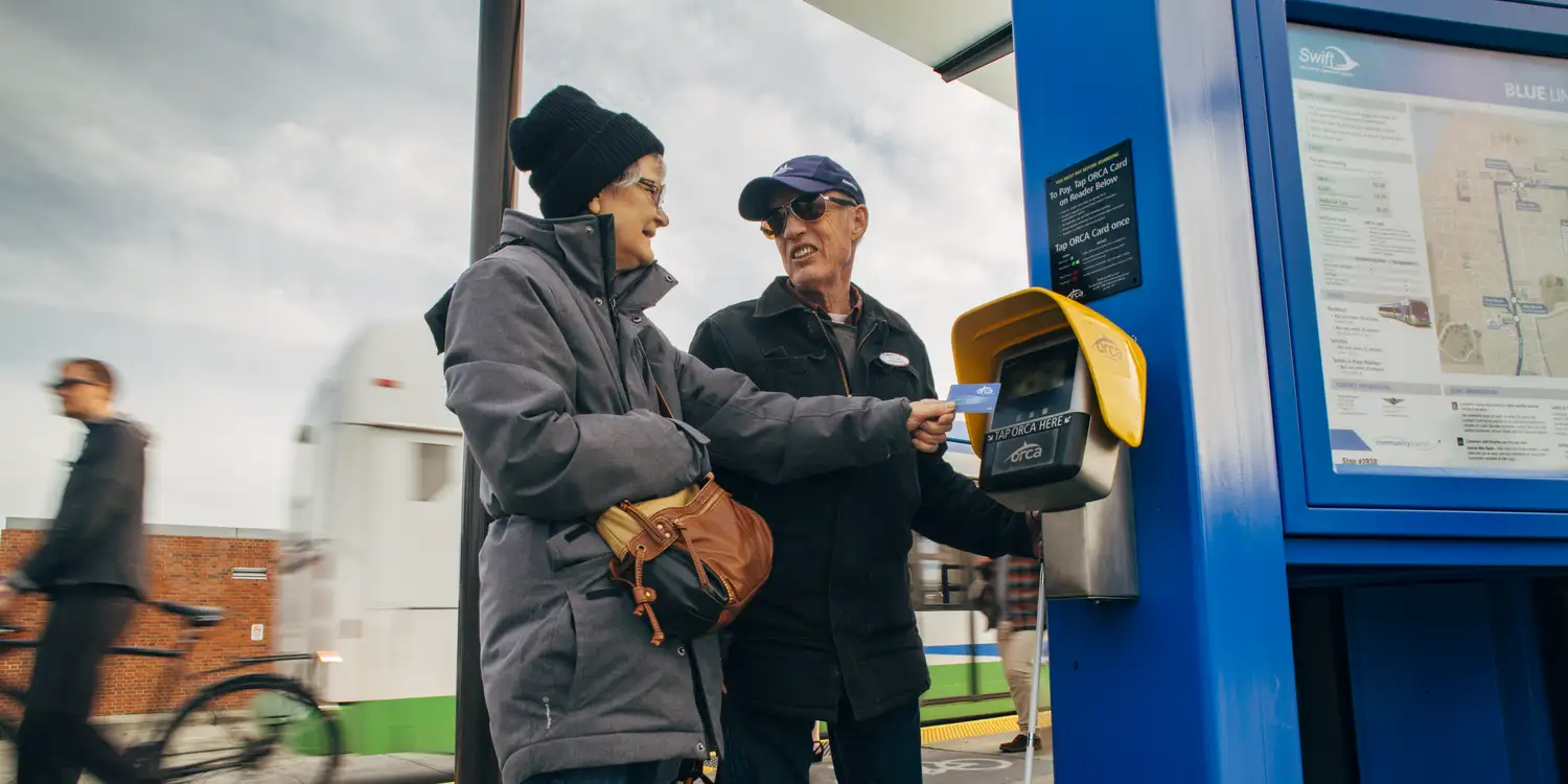 Two riders using an ORCA card reader at a bus stop