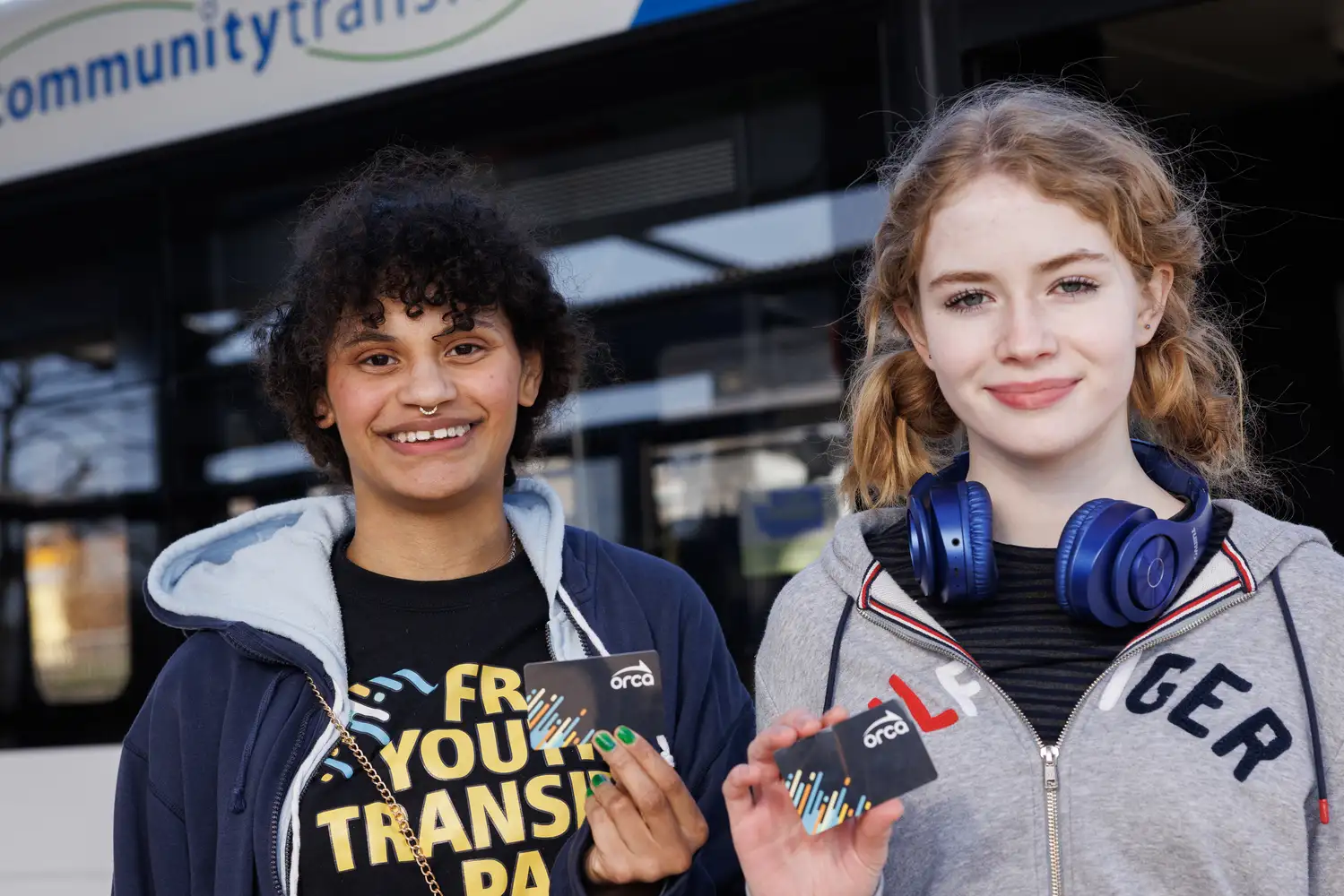 two youth riders holding up their ORCA passes in front of a bus